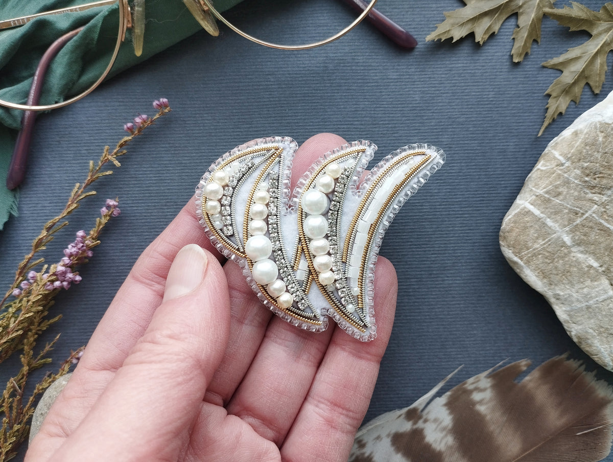 A hand holding a decorative earring with pearls and a floral design against a dark background.
