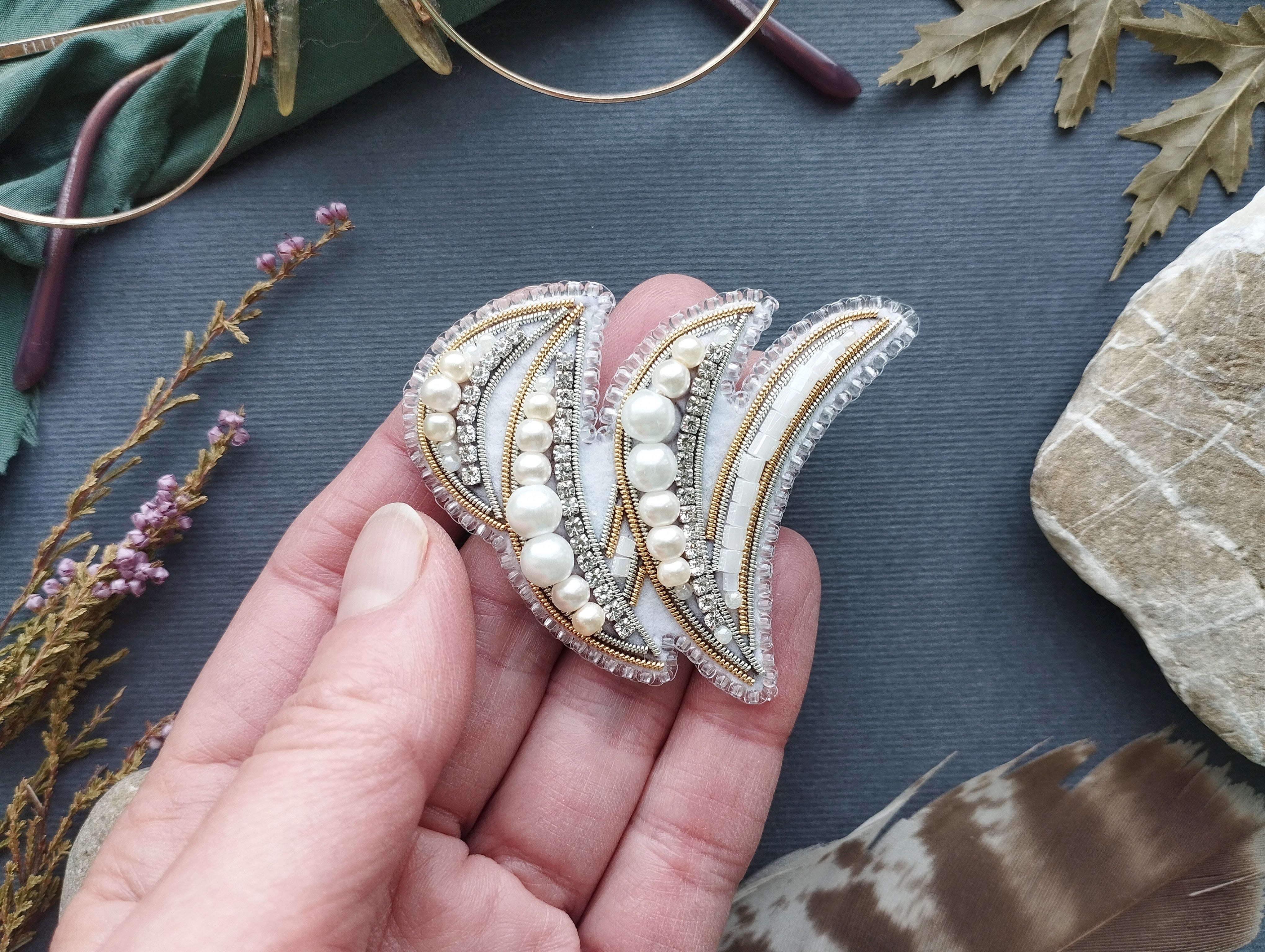A hand holding a decorative earring with pearls and a floral design against a dark background.
