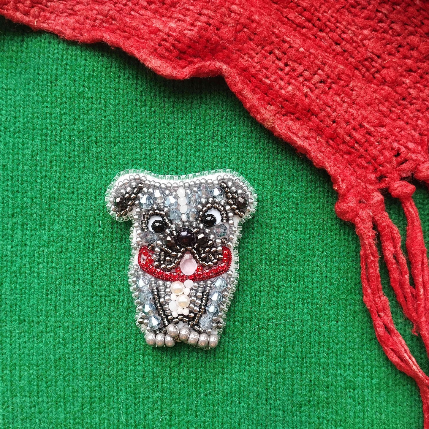 A small, intricately beaded dog brooch is displayed on a green fabric, with a red knitted or crocheted item partially visible to the right.