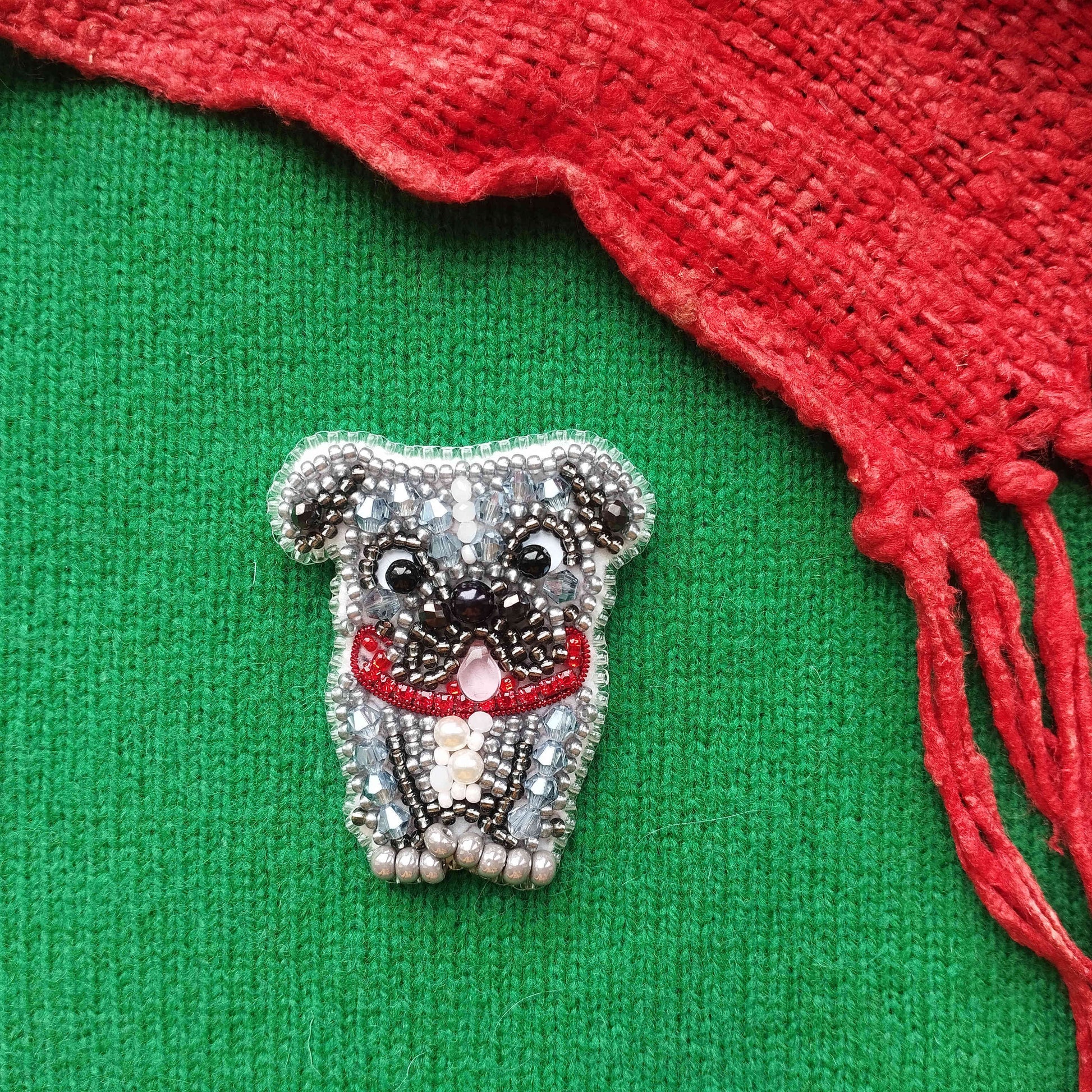 A small, intricately beaded dog brooch is displayed on a green fabric, with a red knitted or crocheted item partially visible to the right.