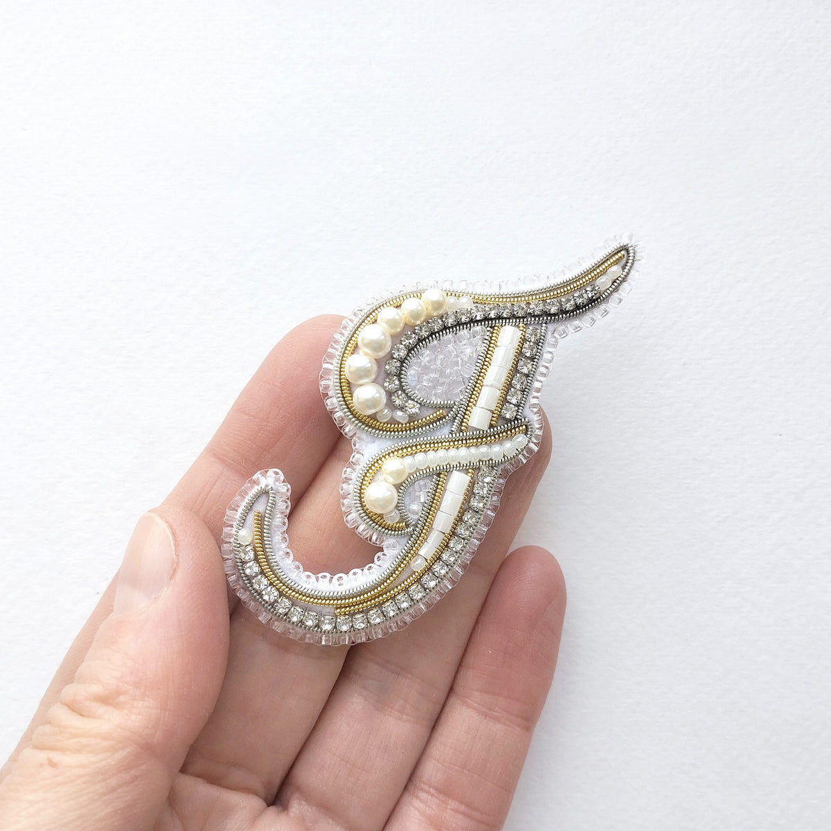 A hand holding a decorative letter 'F' made of beaded and embroidered materials.