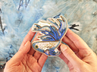 A hand holding a decorative beaded object resembling a fish or sea creature against a blue and white background.