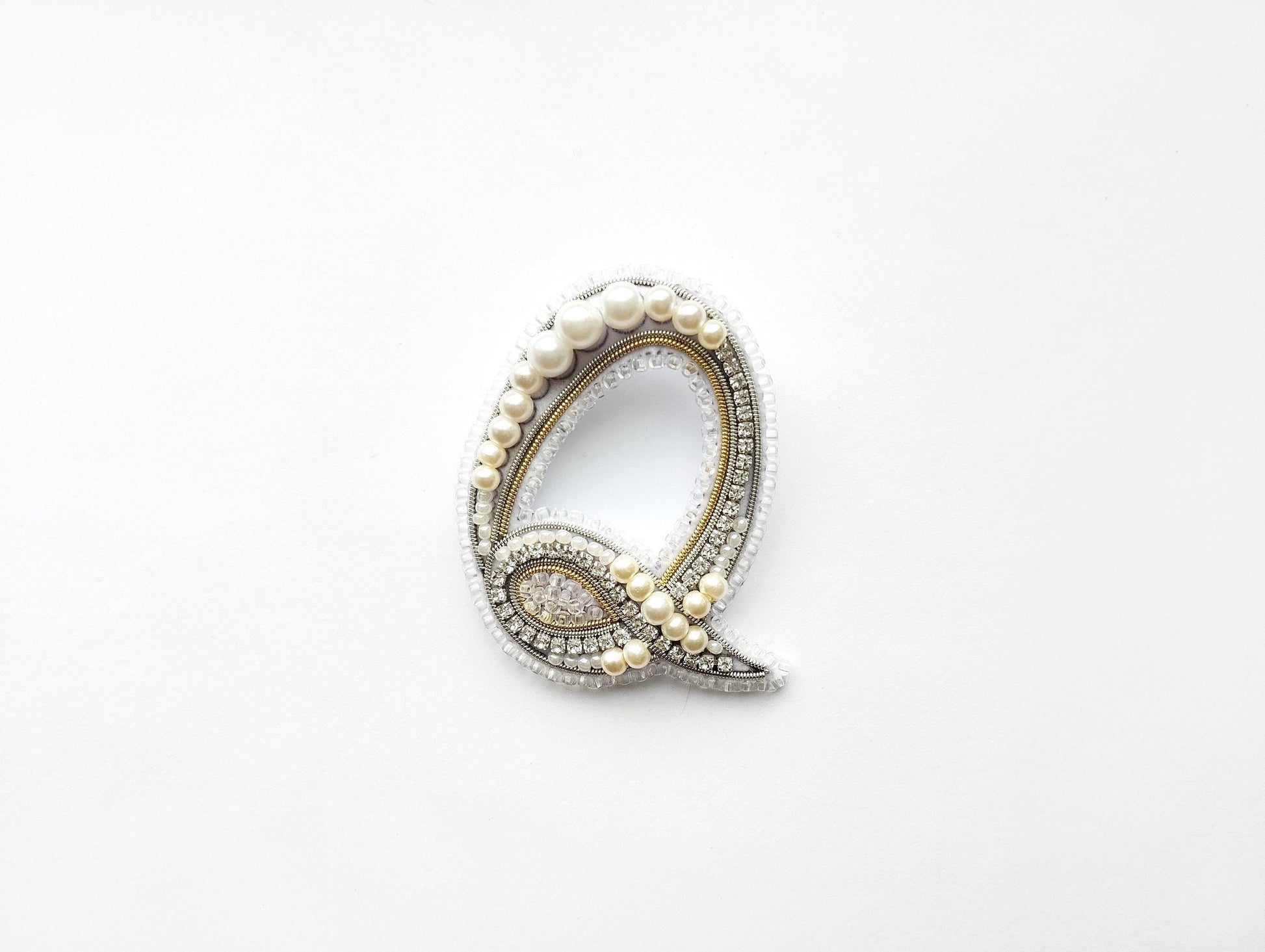 A decorative letter 'Q' made of pearls, diamonds, and other embellishments, displayed against a white background.