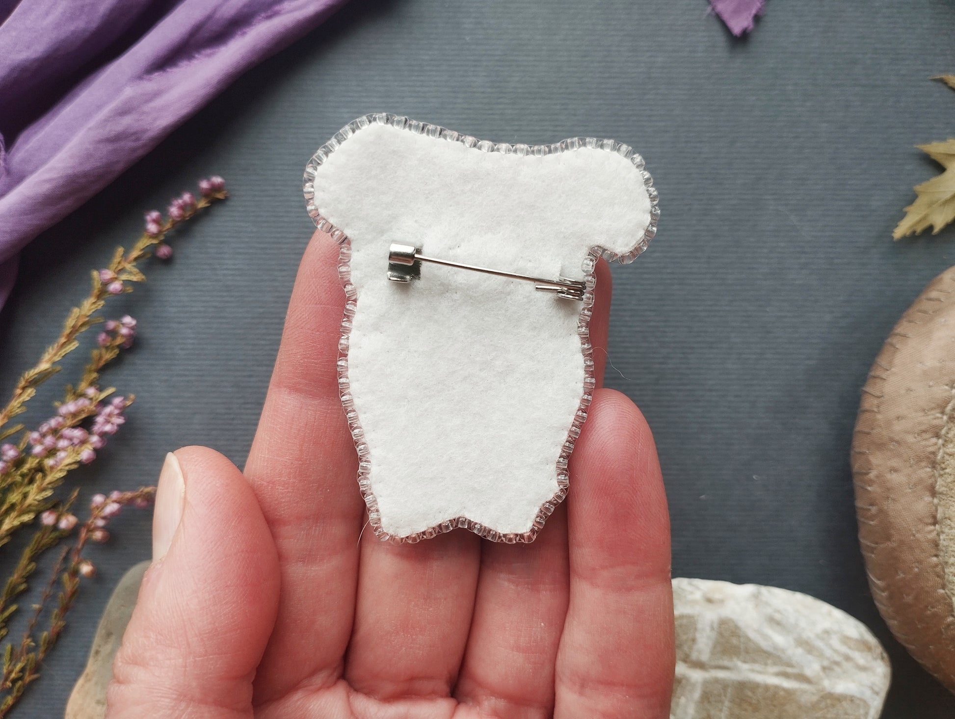 A hand holding a white fabric pin with a silver pinback, surrounded by various objects such as flowers, leaves, and a brown object.