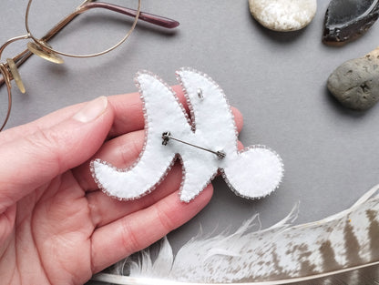 A hand holding a white felt object resembling a crescent moon or star shape, with a pin or pinback attached to it. The hand is positioned over a gray surface, and there are various objects such as glasses, rocks, and feathers scattered around.