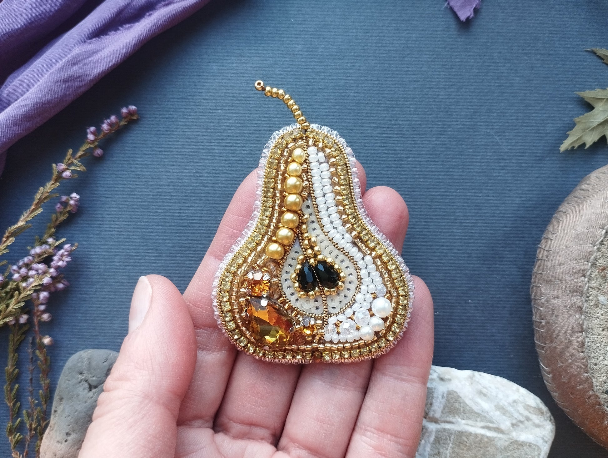 A hand holding a decorative piece of jewelry, which appears to be a beaded pearskin brooch. The jewelry is adorned with various gemstones and pearls, giving it a luxurious and elegant appearance.