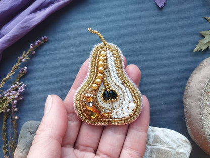 A hand holding a decorative piece of jewelry, which appears to be a beaded pearskin brooch. The jewelry is adorned with various gemstones and pearls, giving it a luxurious and elegant appearance.