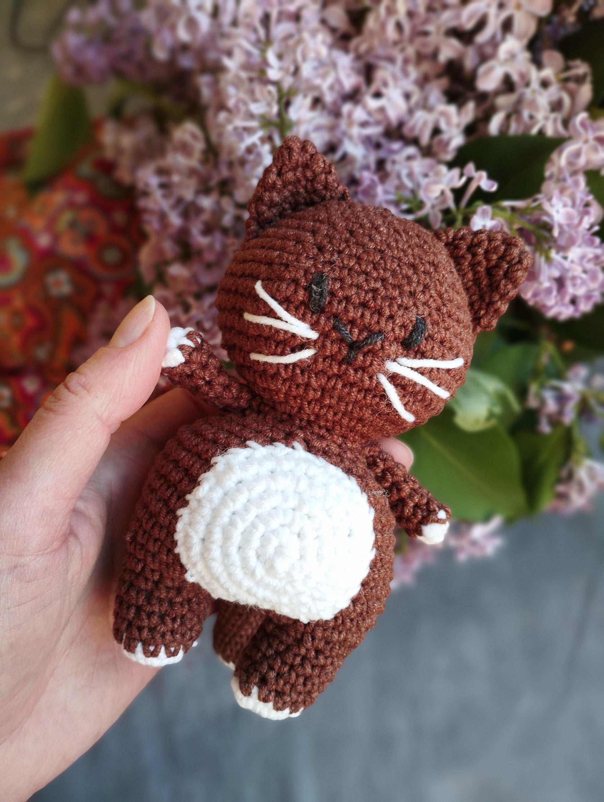 a hand holding a small crocheted cat in front of a bouquet of flowers