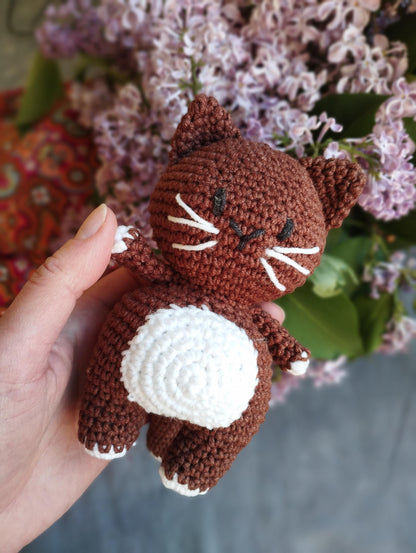a hand holding a small crocheted cat in front of a bouquet of flowers