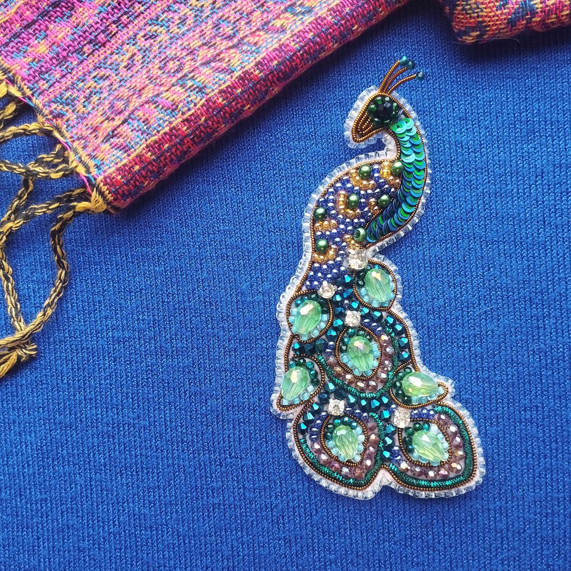 A colorful, intricately designed peacock feather brooch is displayed against a blue background, with a multicolored fabric visible in the background.