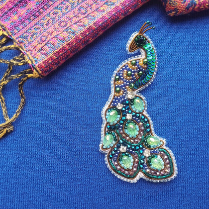 A colorful, intricately designed peacock feather brooch is displayed against a blue background, with a multicolored fabric visible in the background.