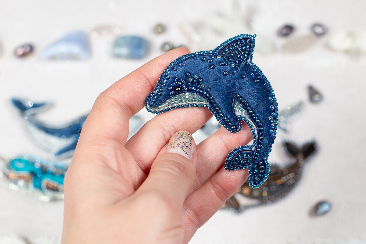 A hand holding a blue, beaded dolphin-shaped object against a background of various other beaded objects.