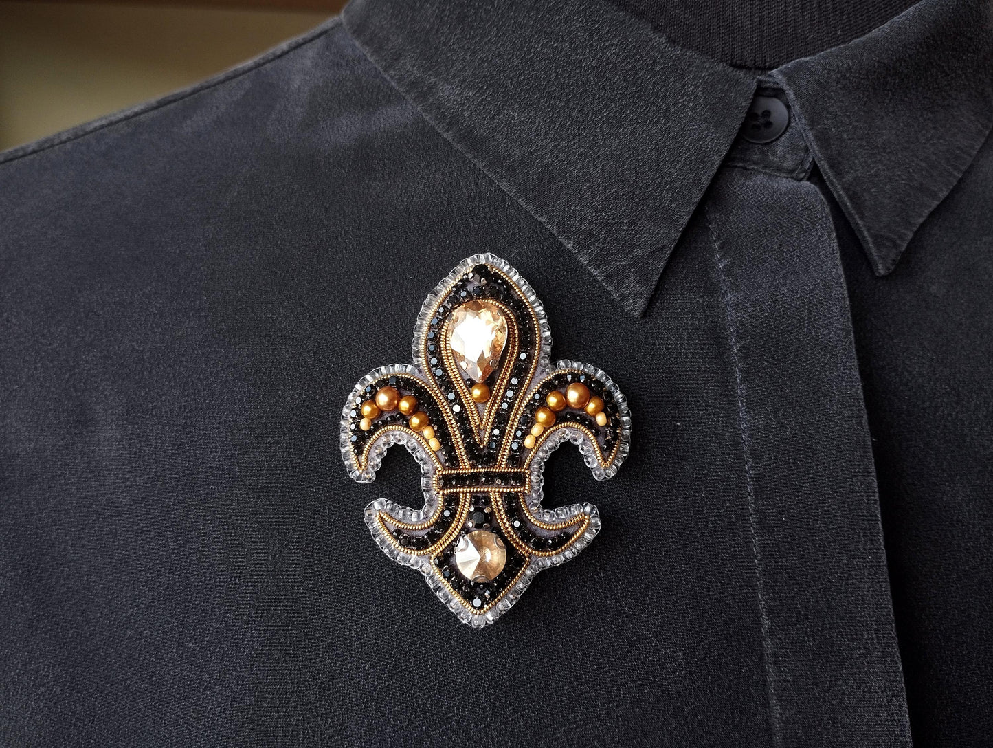 A close-up of a black shirt with a decorative brooch featuring a fleur-de-lis design and gold and black embellishments.