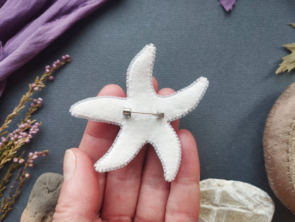 A hand holding a white felt starfish with a pin in the center, surrounded by various natural elements such as leaves, flowers, and rocks.