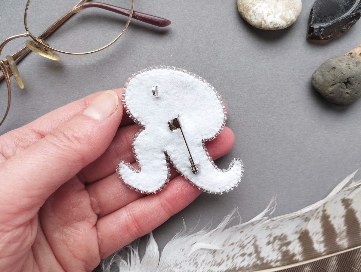 A hand holding a white felt octopus pin, surrounded by various objects such as glasses, rocks, and feathers on a gray background.