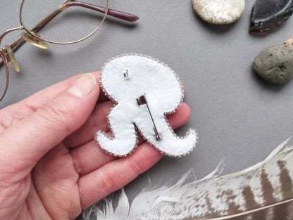 A hand holding a white felt octopus pin, surrounded by various objects such as glasses, rocks, and feathers on a gray background.