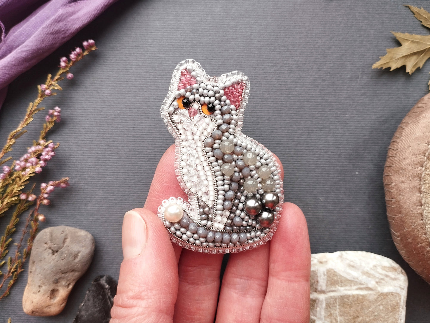 A hand is holding a decorative beaded cat pin against a gray background.