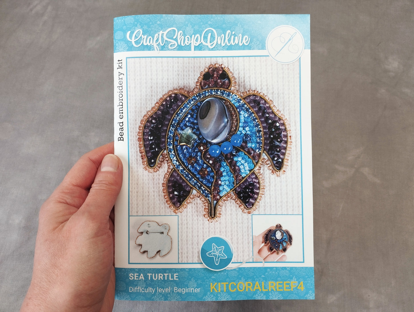 A hand is holding a kit for creating a sea turtle embroidery, which includes a detailed image of the completed piece and instructions for the embroidery process.