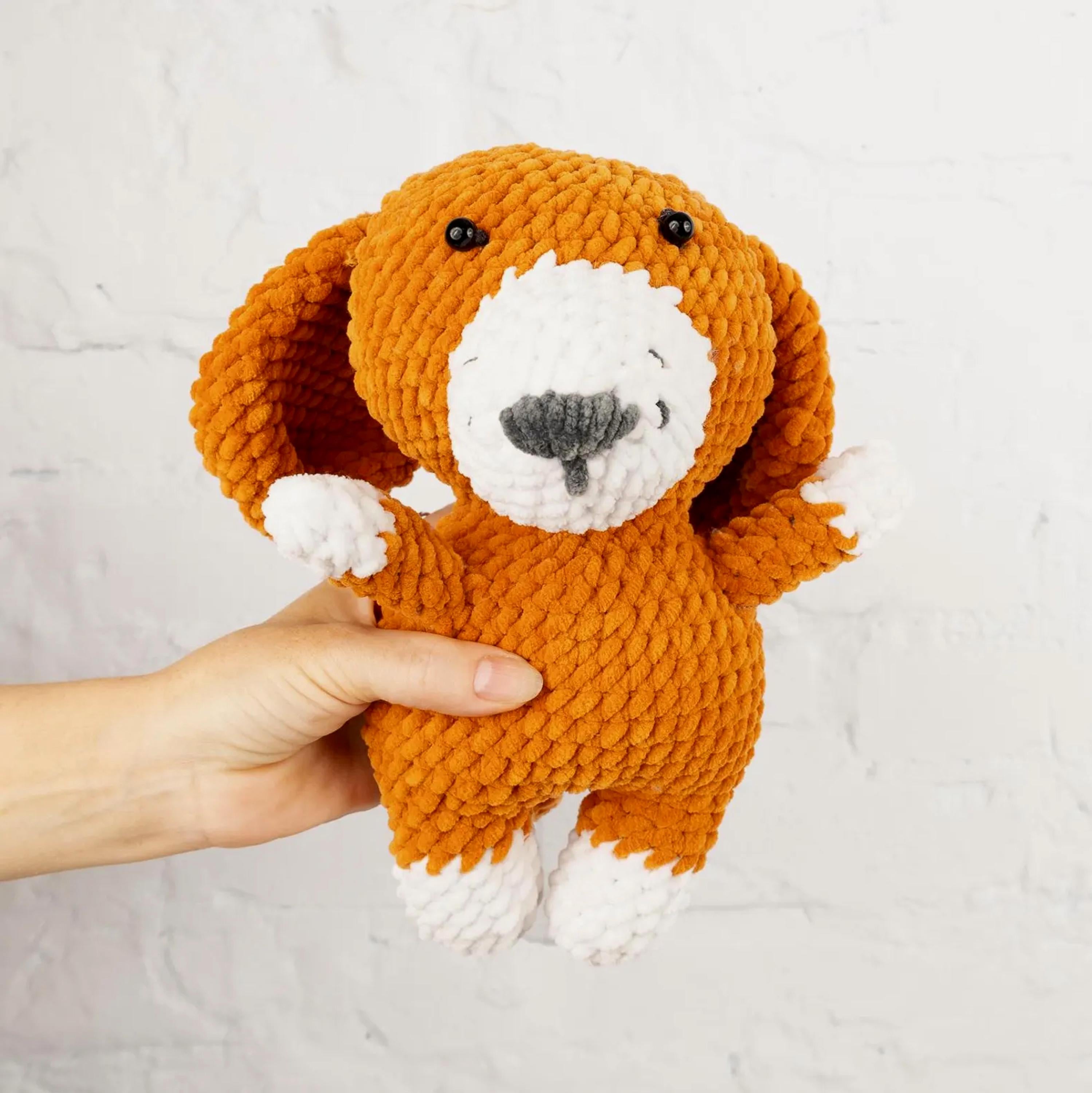 A hand is holding a brown and white crocheted stuffed dog toy against a white background.