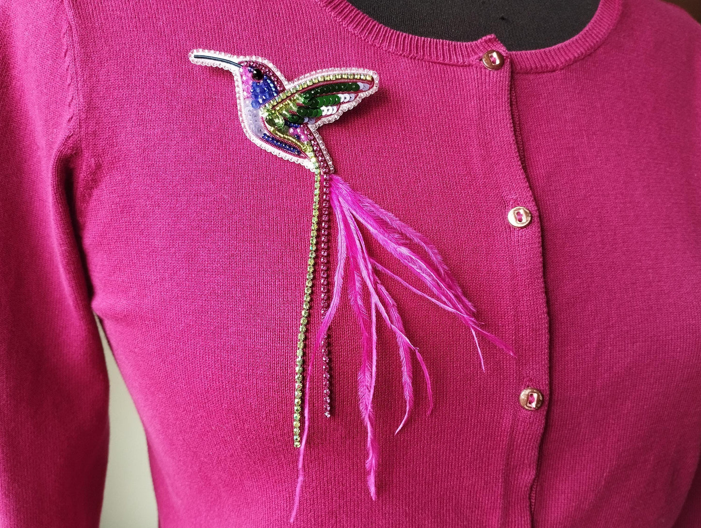 A close-up of a pink sweater with a colorful, beaded hummingbird brooch on the front.