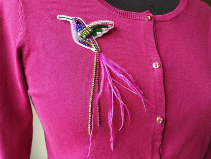 A close-up of a pink sweater with a colorful, beaded hummingbird brooch on the front.
