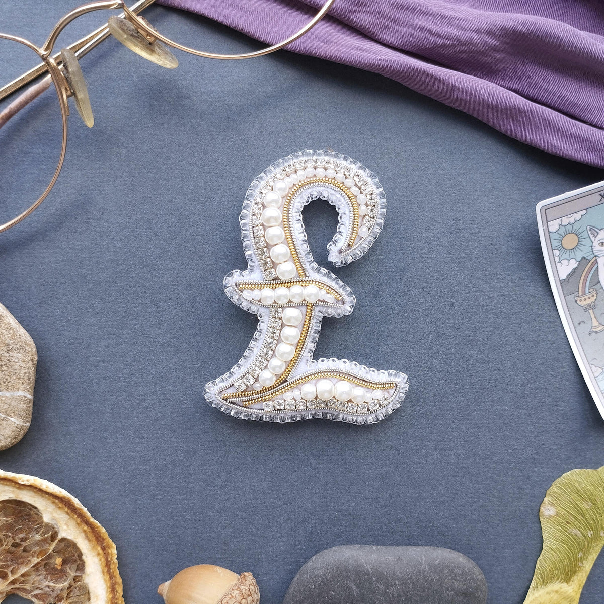 a close-up view of a decorative monogram made of pearls and beads, surrounded by various objects such as a pair of glasses, a tarot card, and some natural elements like an orange and a rock.