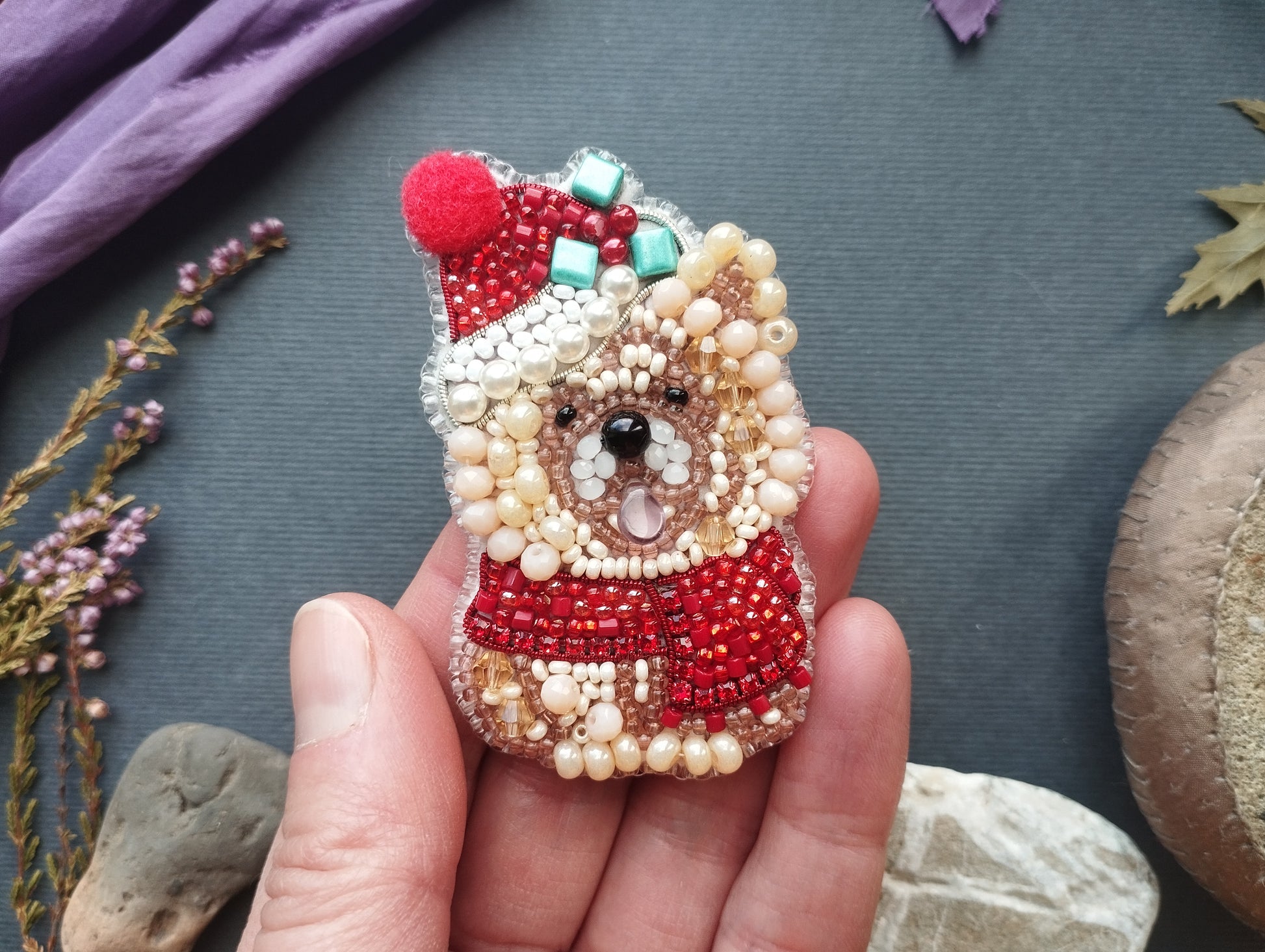A hand holding a small, intricately beaded pin or brooch in the shape of a dog wearing a Santa hat and scarf.
