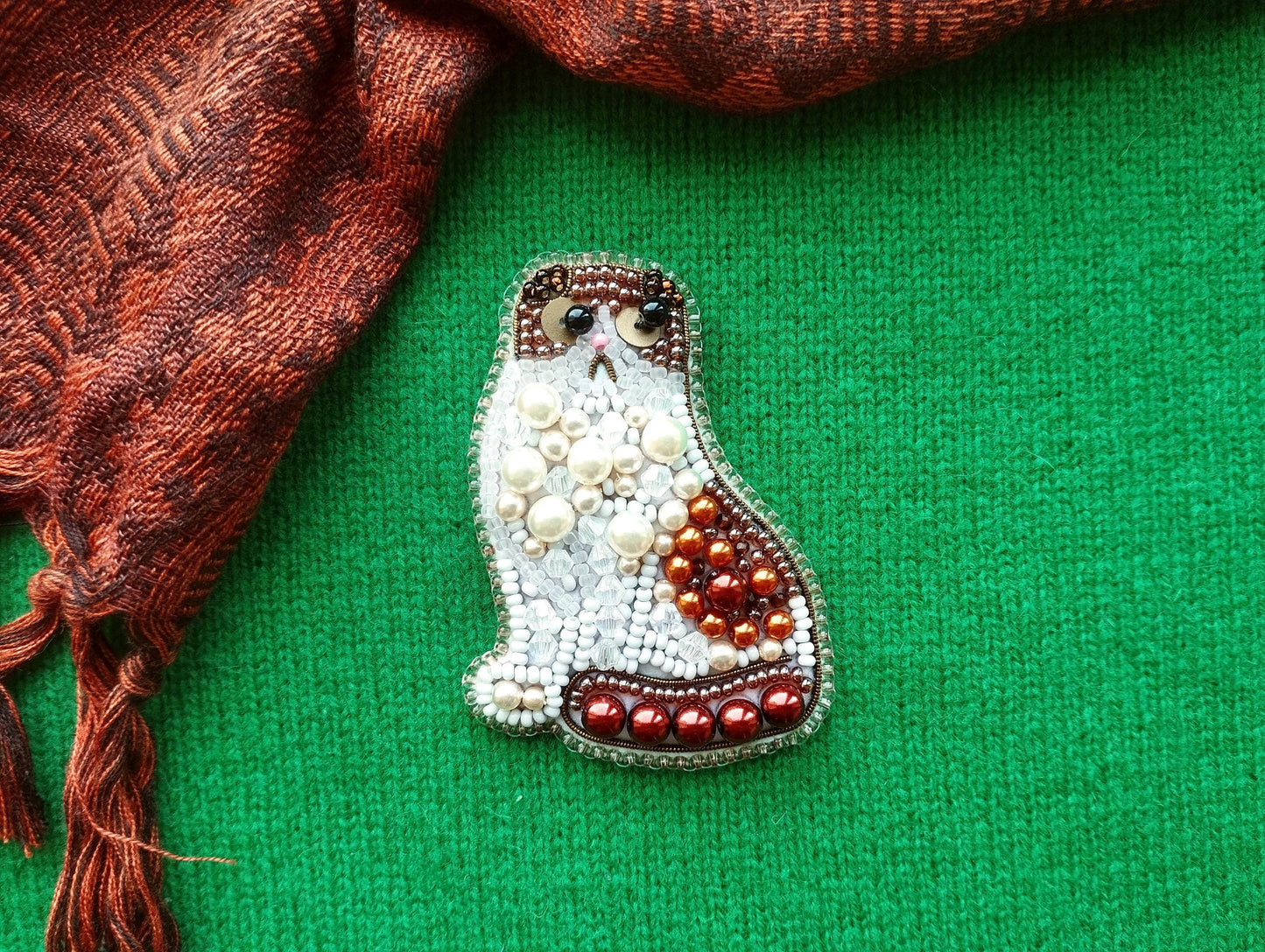 A decorative cat brooch made of beads and pearls is displayed against a green background, with a red and brown patterned fabric visible on the left side.