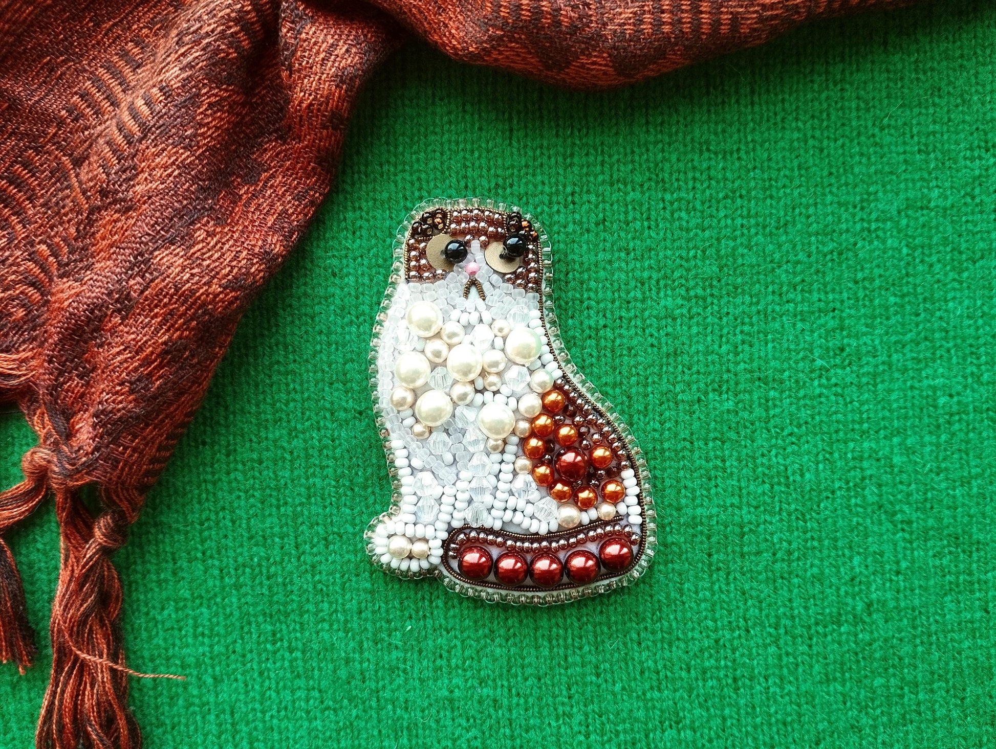 A decorative cat brooch made of beads and pearls is displayed against a green background, with a red and brown patterned fabric visible on the left side.