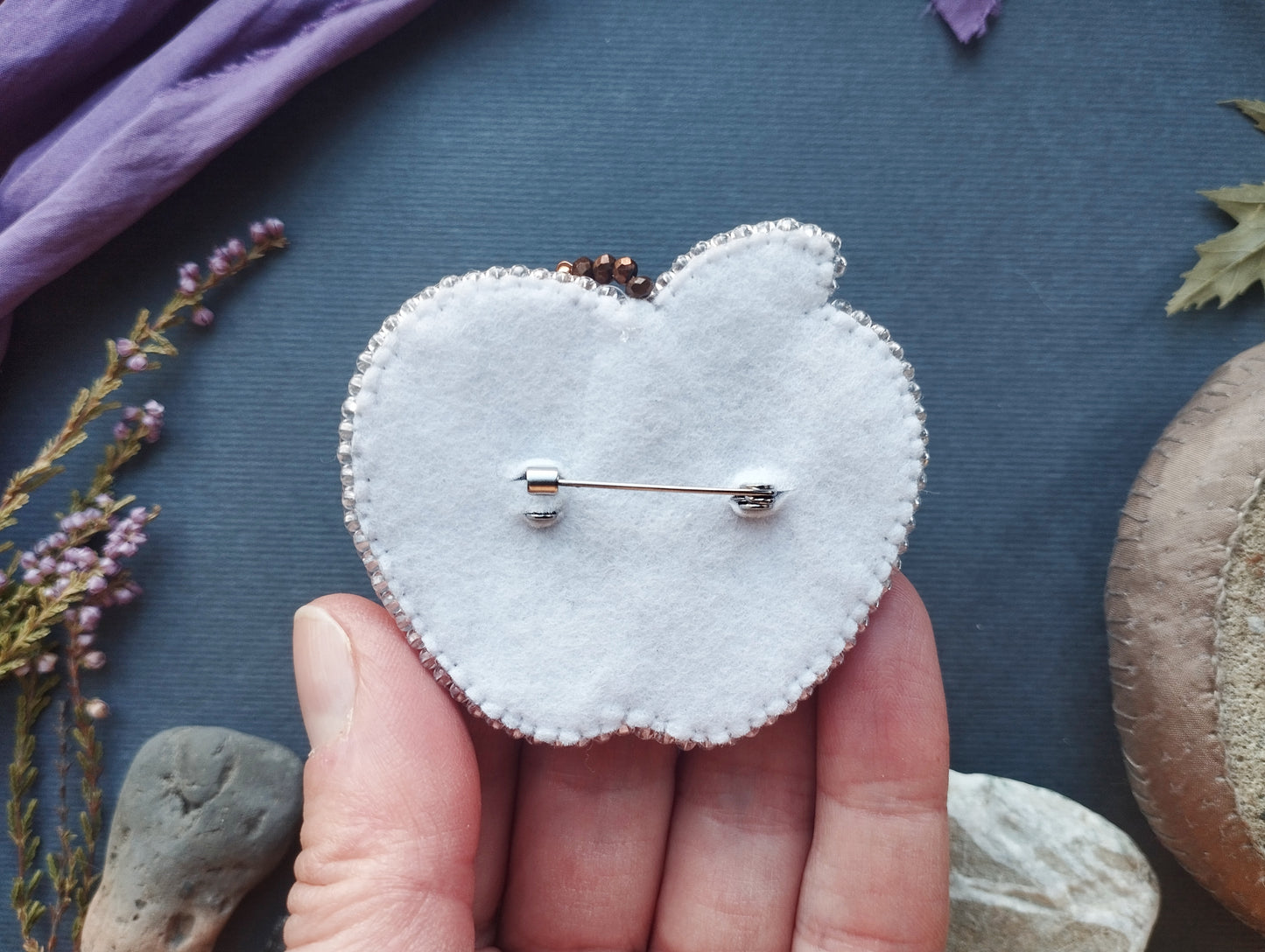 A hand holding a white felt apple pin against a dark background with various objects, including flowers and rocks.
