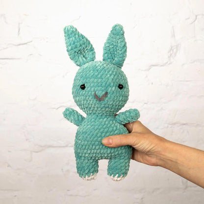 A hand is holding a small, crocheted green bunny toy against a white brick wall background.