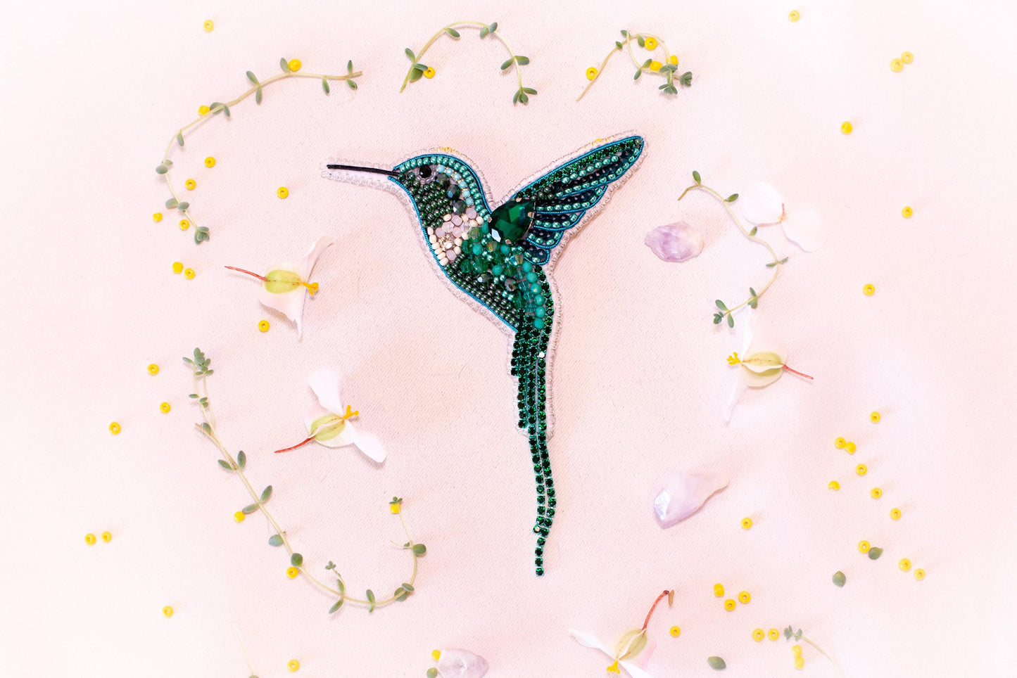 a close-up of a green hummingbird brooch adorned with rhinestones, surrounded by yellow flowers and green leaves.