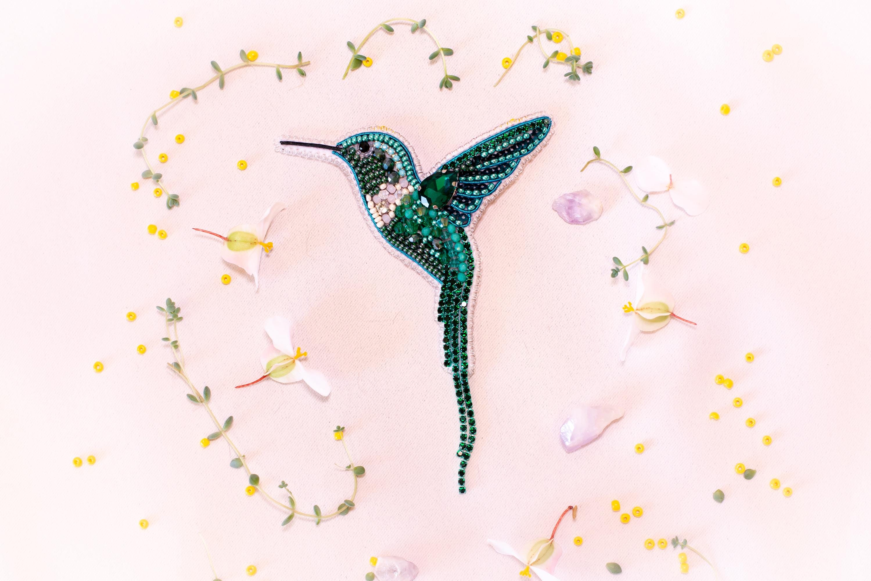 a close-up of a green hummingbird brooch adorned with rhinestones, surrounded by yellow flowers and green leaves.