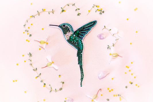 a close-up of a green hummingbird brooch adorned with rhinestones, surrounded by yellow flowers and green leaves.