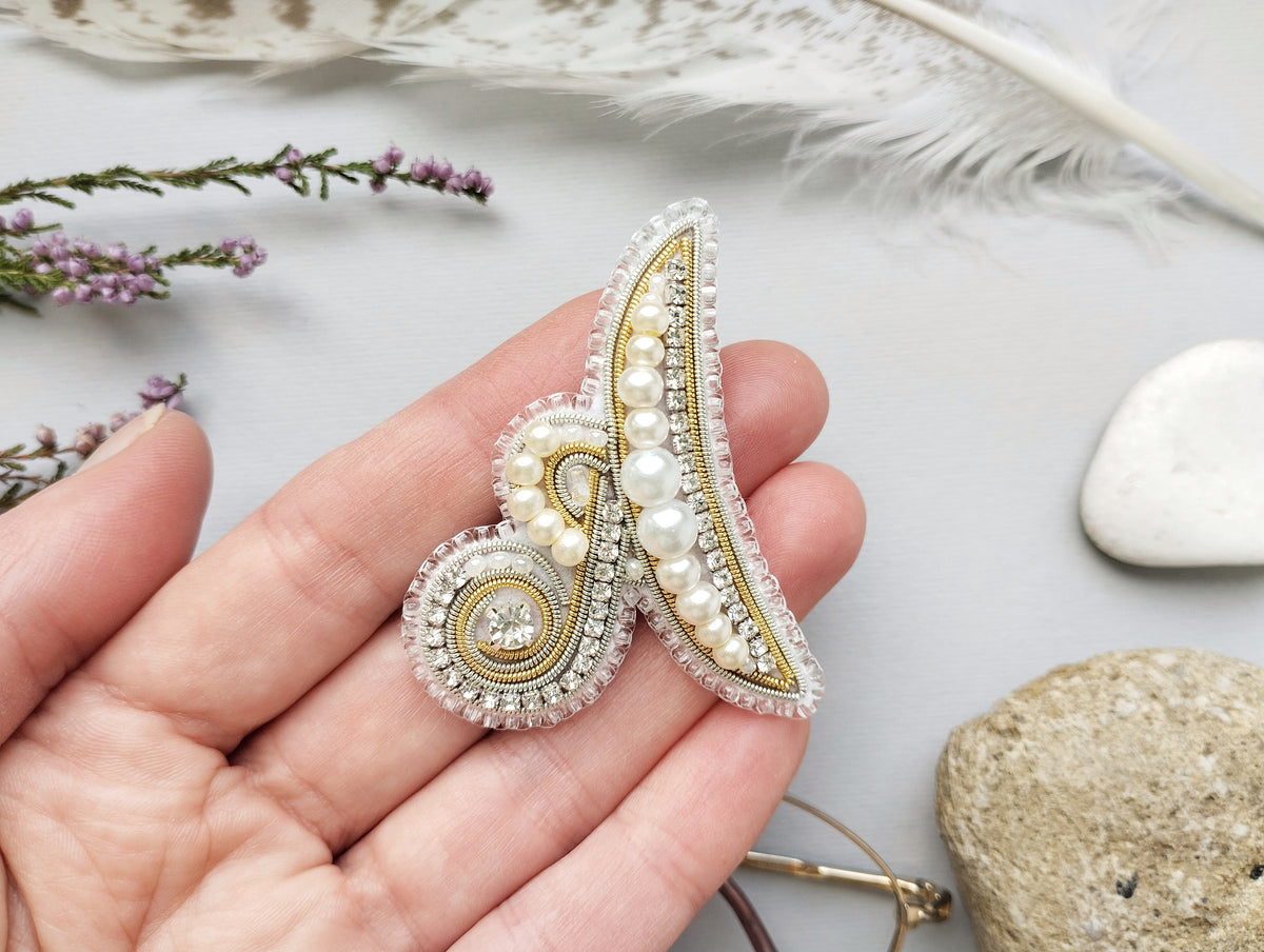 A hand holding a decorative piece of jewelry, which appears to be a brooch or pin, with a combination of pearls, beads, and a gemstone. The background includes feathers, rocks, and flowers, creating a natural and artistic setting.