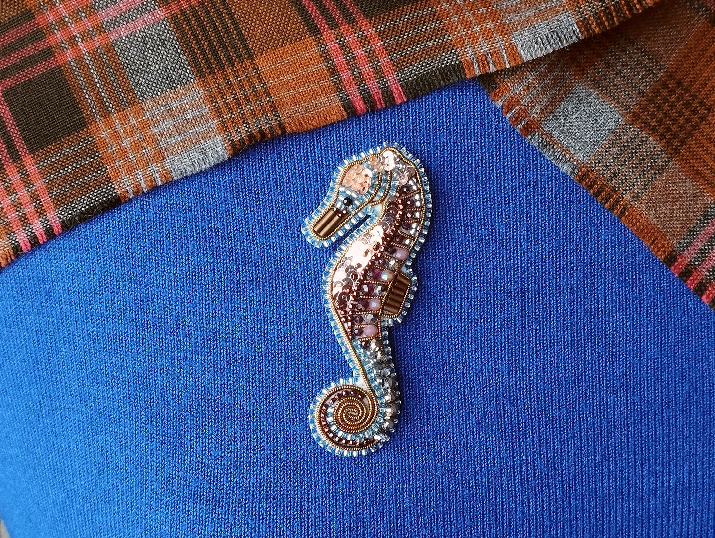 A decorative beaded seahorse brooch is displayed on a blue fabric surface, with a plaid fabric visible in the background.