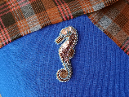 A decorative beaded seahorse brooch is displayed on a blue fabric surface, with a plaid fabric visible in the background.