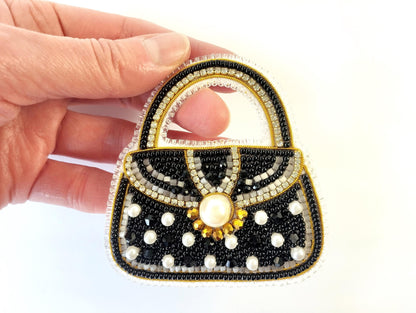 A hand holding a handbag-shaped embroidered piece with black, white, and gold beads and a pearl at the center.