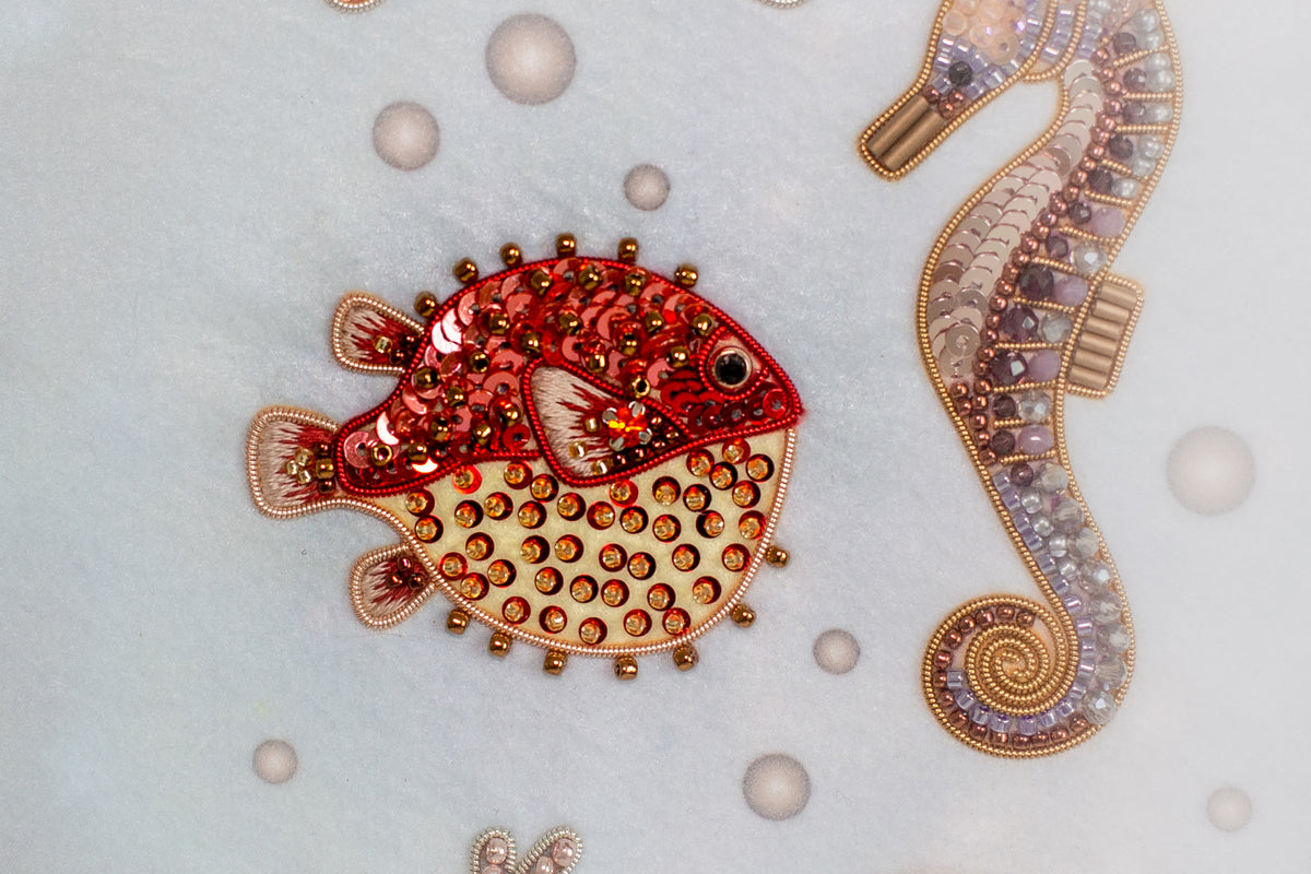 a detailed embroidered design of a red and yellow fish with gold and silver embellishments, surrounded by bubbles and a purple and gold seahorse.