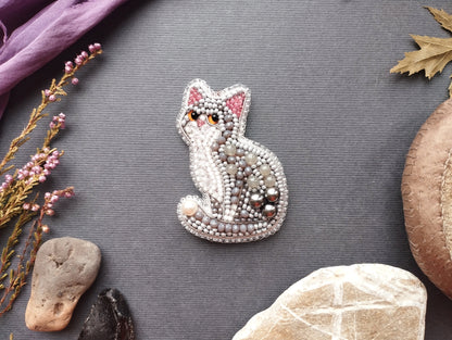 a decorative cat brooch made of beads and pearls, surrounded by various natural elements such as leaves, rocks, and flowers on a grey background.