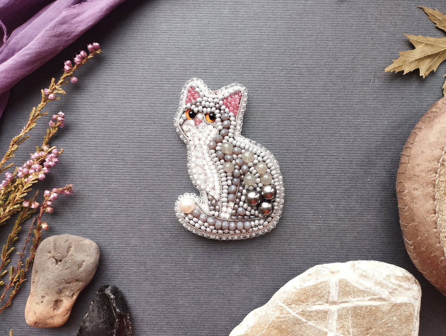 a decorative cat brooch made of beads and pearls, surrounded by various natural elements such as leaves, rocks, and flowers on a grey background.