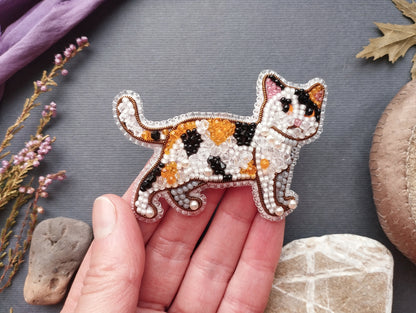 A hand holding a beaded embroidered cat pin against a gray background with various natural elements, such as flowers and rocks.