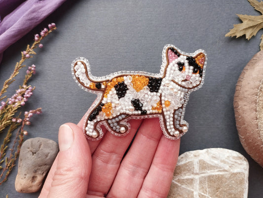 A hand holding a beaded embroidered cat pin against a gray background with various natural elements, such as flowers and rocks.