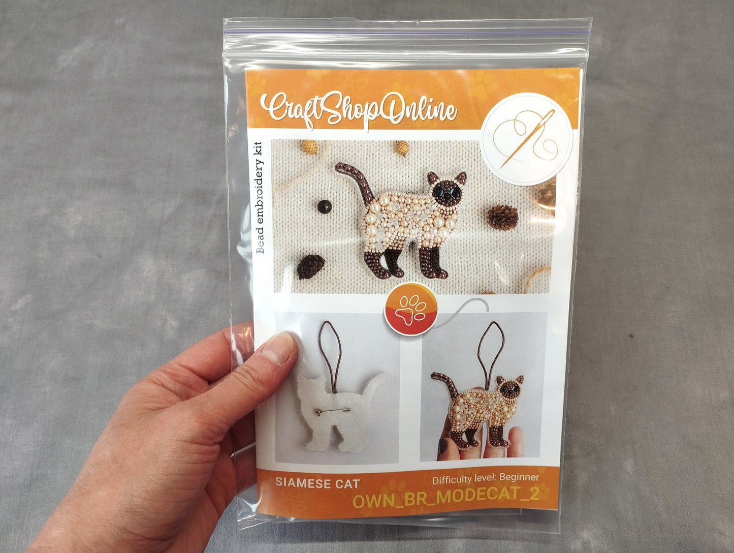 A hand holding a package containing a kit for creating embroidered cat ornaments.