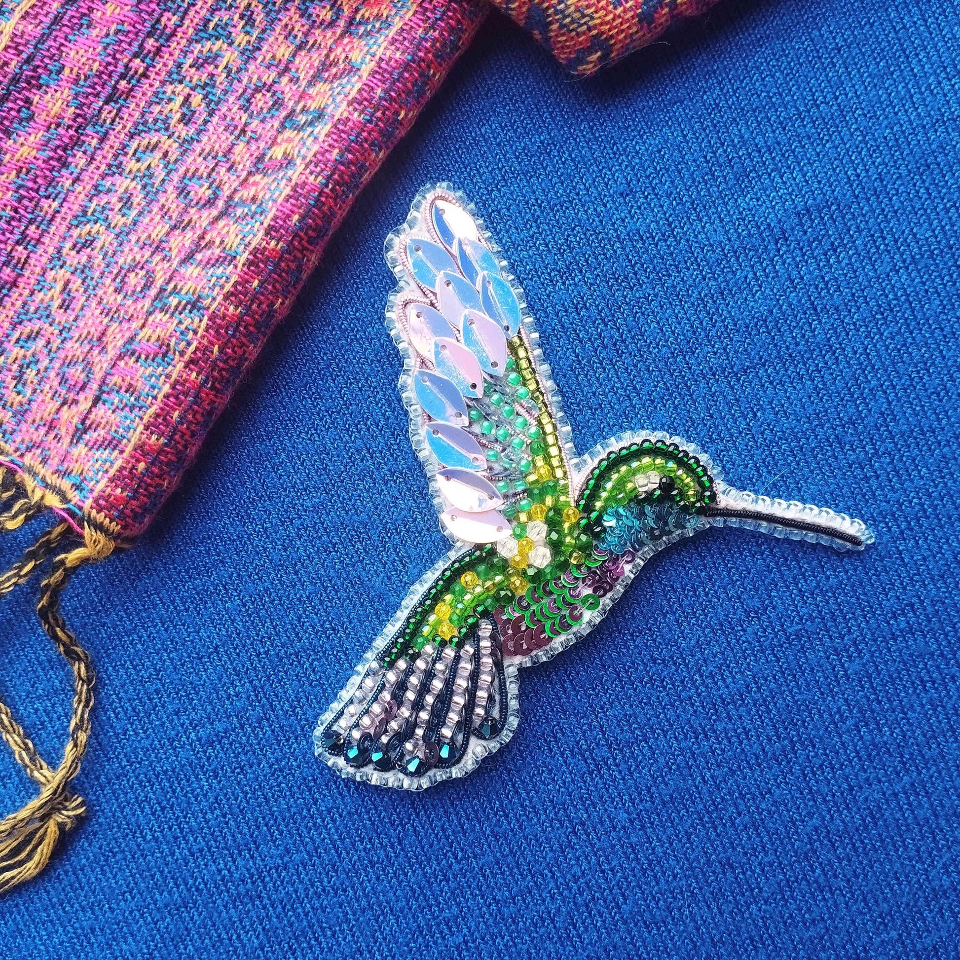A colorful, intricately designed hummingbird brooch is displayed on a blue fabric surface, with a multicolored scarf partially visible in the background.