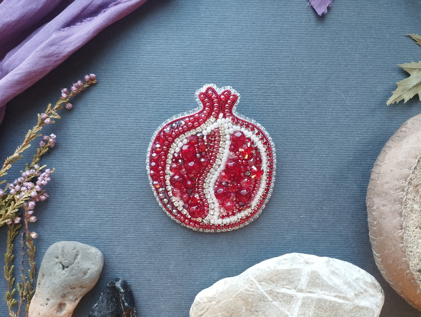 a pomegranate-shaped brooch adorned with red and white beads, surrounded by various natural elements such as rocks, leaves, and flowers on a blue background.