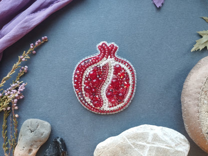 a pomegranate-shaped brooch adorned with red and white beads, surrounded by various natural elements such as rocks, leaves, and flowers on a blue background.