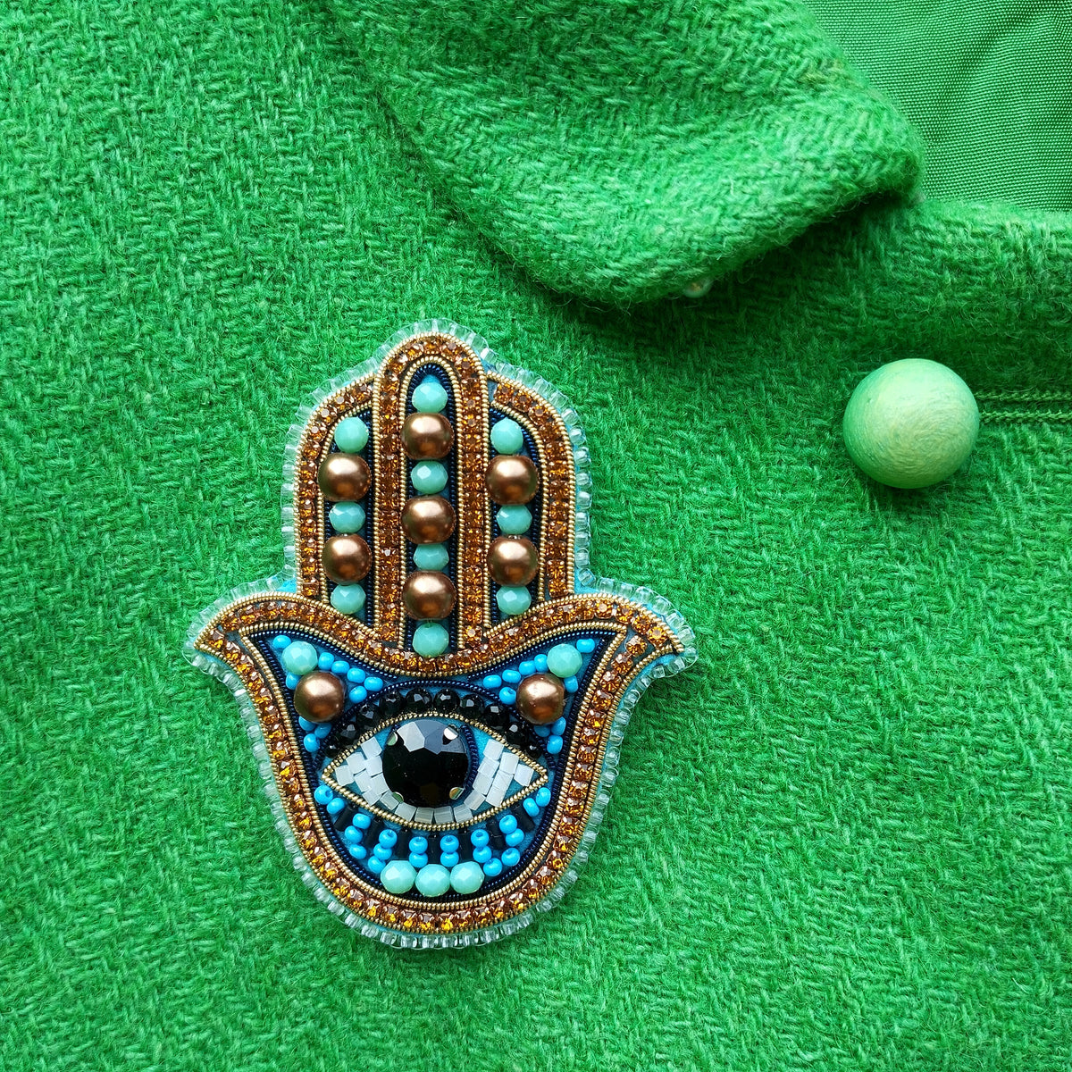 a close-up of a handcrafted brooch with a blue and gold design, adorned with a large eye and surrounded by small beads. The brooch is placed on a green fabric, which provides a contrasting background.