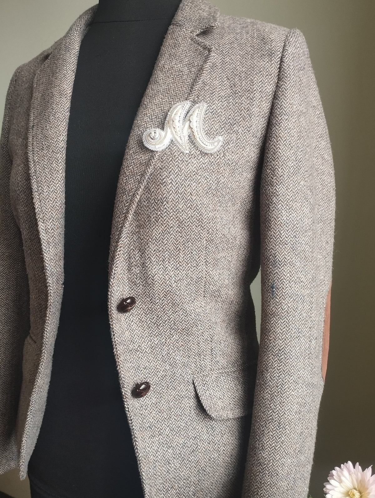 A gray tweed blazer with a white embroidered letter "M" on the lapel, displayed on a mannequin.