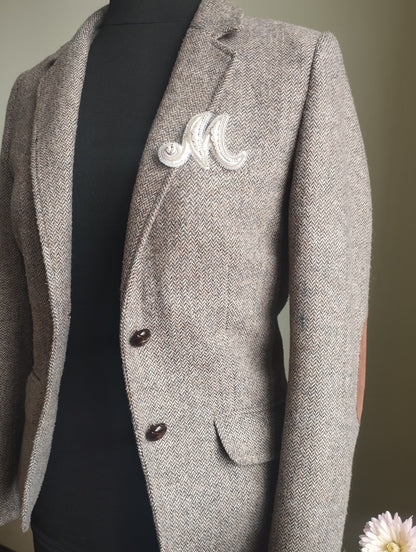 A gray tweed blazer with a white embroidered letter "M" on the lapel, displayed on a mannequin.